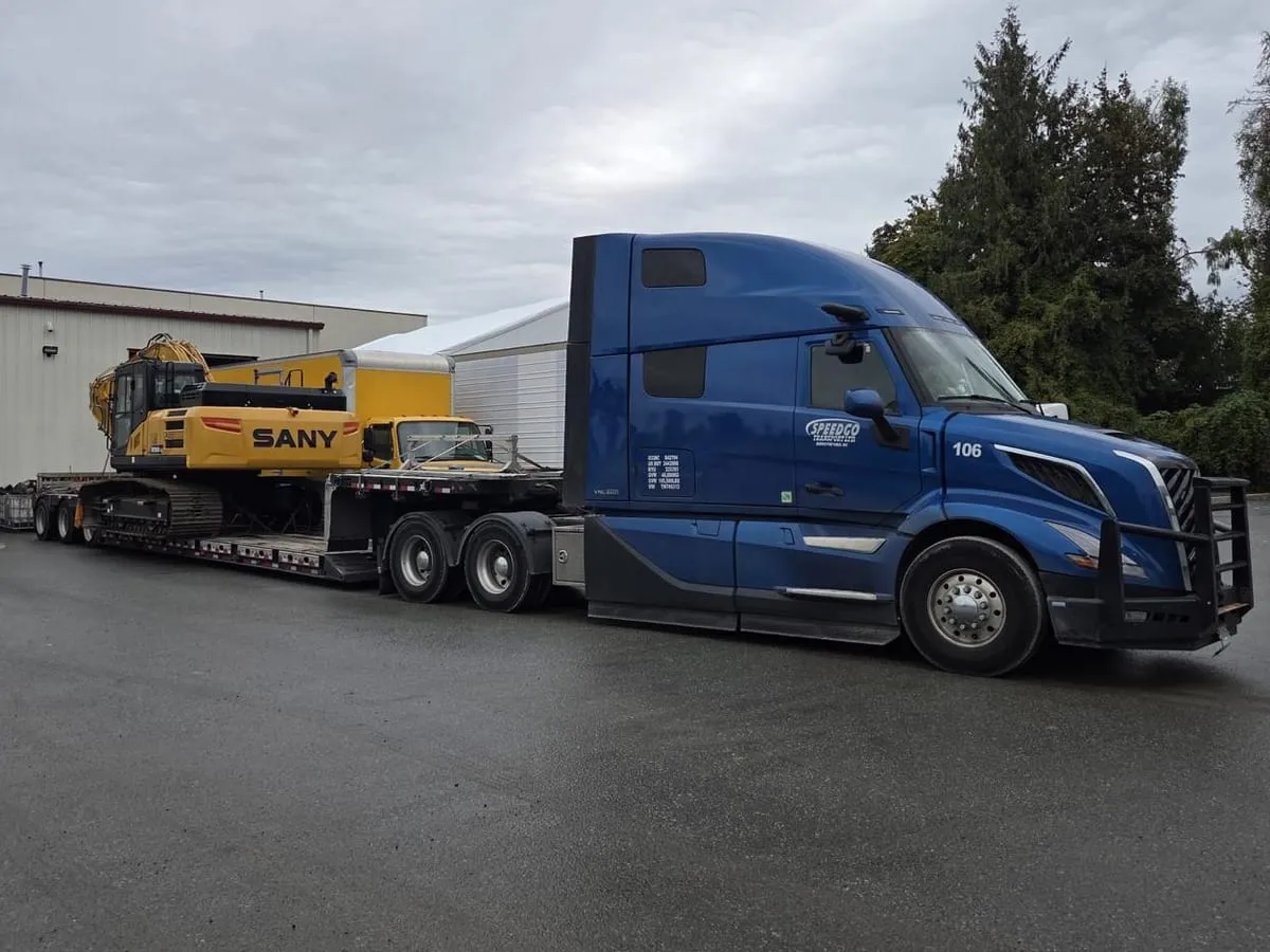 Blue Speedgo Transport truck hauling a SANY excavator, parked outside a warehouse