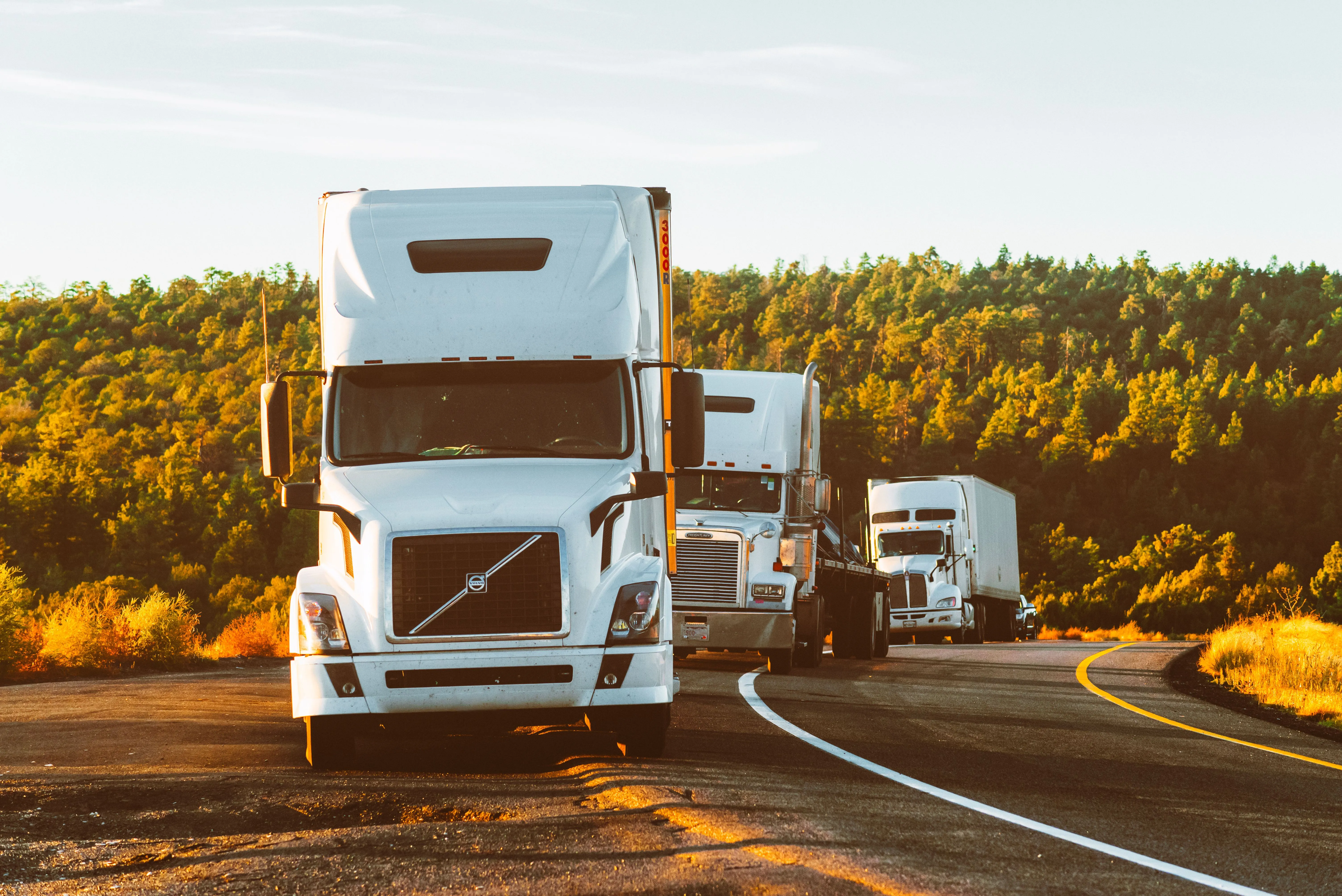 A line of transport trucks from partner carriers hauling freight along a highway at sunset