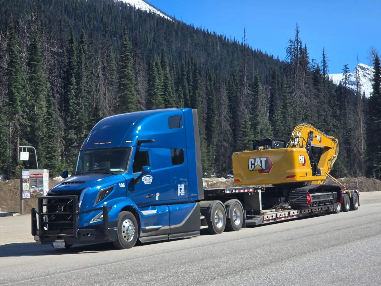 Blue Speedgo Transport Volvo truck hauling a yellow CAT excavator on a 53' RGN trailer through the mountains