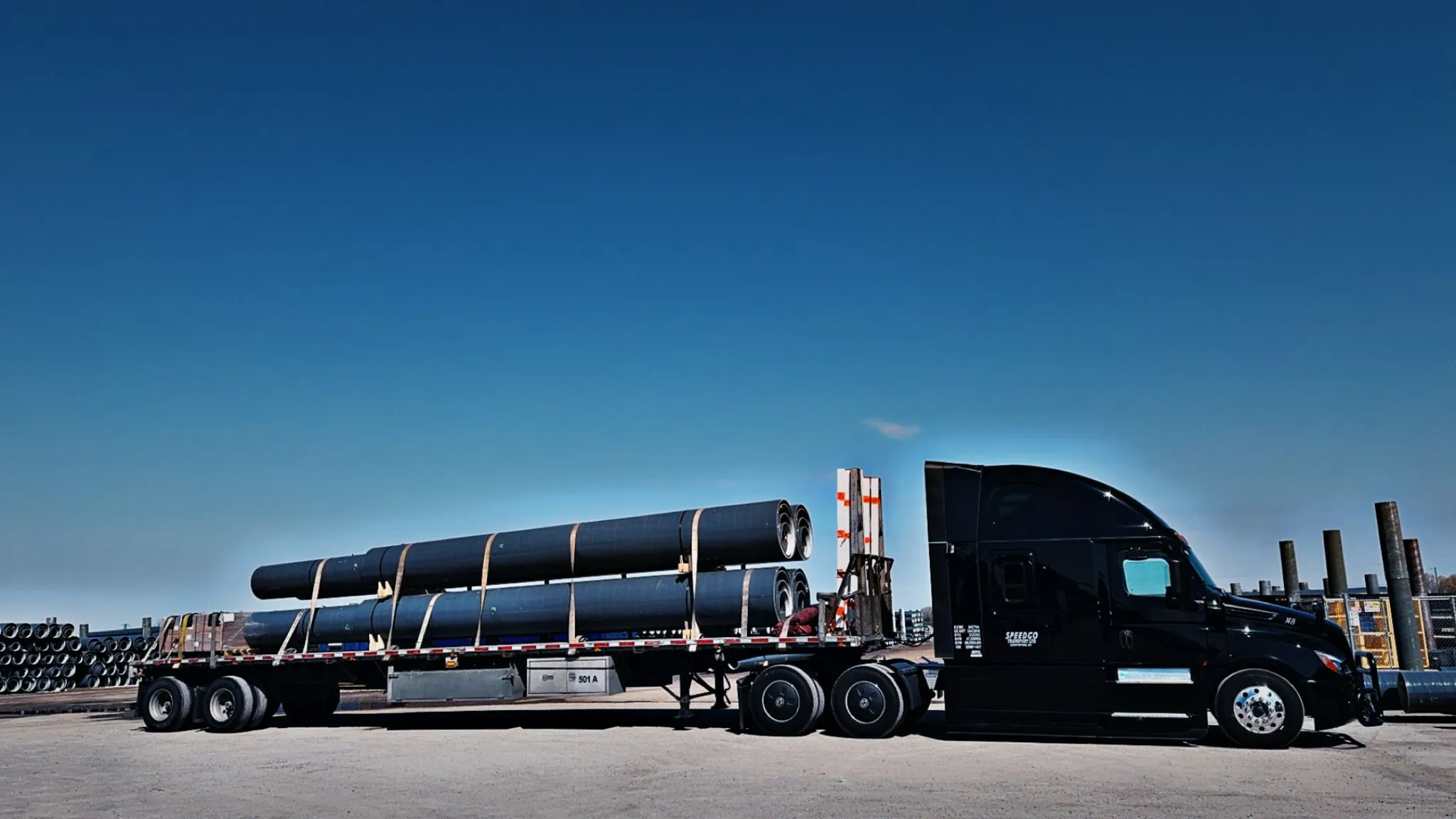Black Speedgo Transport truck hauling large steel pipe on a tandem flat deck trailer